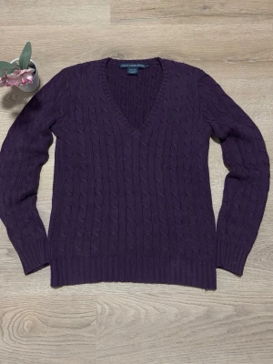 Lila kabelstickad tröja Polo Ralph Lauren - Snygg lila kabelstickad tröja från Polo Ralph Lauren med djup v-ringning. Tröjan har långa ärmar och ribbade muddar vid ärmslut och nederkant. Perfekt för dig som gillar klassisk och tidlös stil.