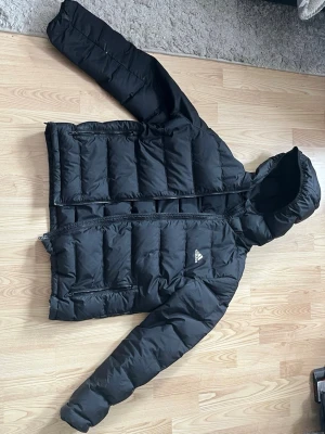 Svart pufferjacka från Adidas - Säljer en svart pufferjacka från Adidas med huva och dragkedja framtill. Jackan har två stora inner fickor ocj två sidofickor och med dragkedja och Adidas-logga på bröstet. Perfekt för kalla vinterdagar tack vare den tjocka vadderingen och den klassiska pufferlooken. storlek large men passar bra om man har medium också.