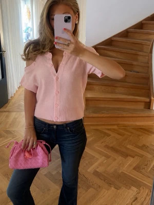 Ljusrosa stickad topp med knappar - Superfin ljusrosa stickad topp med korta ärmar och v-ringning. Toppen har knappar framtill och en offshoulder-design som ger en trendig look. Perfekt att matcha med jeans eller kjol för en söt och avslappnad stil.