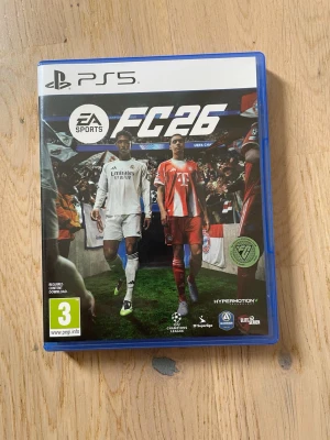 EA Sports FC 26 till PS5 - Säljer spelet EA Sports FC 26 till PlayStation 5. Spelet kommer i ett blått plastfodral med omslag som visar två fotbollsspelare på en gräsplan. Skivan är svart, vit och grön med spelets logga. Perfekt för dig som gillar fotbollsspel och vill spela Champions League.