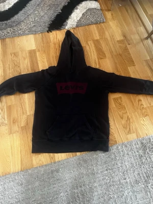Svart hoodie från Levi's med logga - Säljer en svart hoodie från Levi's med stor röd logga på bröstet. Hoodien har en klassisk känguruficka framtill och en stor huva. Tillverkad i mjukt material som passar perfekt för en avslappnad stil.