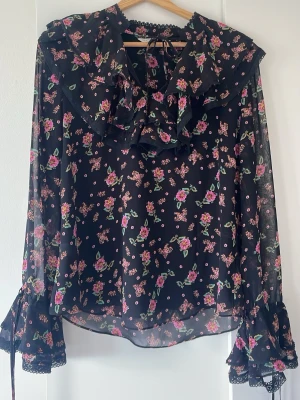 Svart blommig volangblus med spets från Rebecka Minkoff - Romantisk svart blus med rosa och gröna blommor, volangkrage och knytning i nacken. Blusen är en Rebecka Minkoff och såldes slut snabbt när den kom ut! Långa, transparenta ärmar med spetsdetaljer och volanger vid ärmslut. Tillverkad i ett tunt, luftigt material som ger en bohemisk känsla. 
