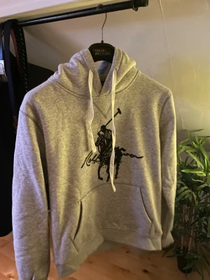 Grå hoodie med polo-tryck - Snygg grå hoodie med stort svart polo-tryck på bröstet. Klassisk känguruficka framtill och ribbade muddar. Perfekt för dig som gillar sportiga och stilrena plagg. Materialet är mjukt och bekvämt, passar till chill och streetwear.