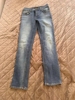 Blå raka jeans från Nudie Jeans - Säljer ett par klassiska blå jeans från Nudie Jeans med raka ben och snygg slitning på låren. Jeansen har fem fickor, bälteshällor och kontrastsömmar. Perfekta till en avslappnad stil och tillverkade i mjuk denim.