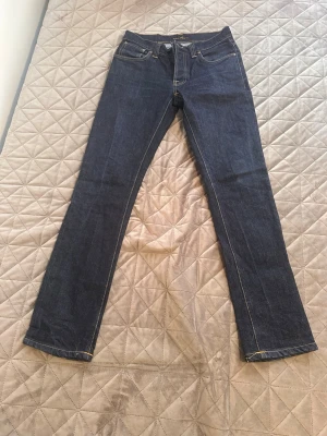 Mörkblå jeans från Nudie Jeans - Säljer ett par mörkblå jeans från Nudie Jeans med raka ben och klassisk femficksmodell. Jeansen har kontrastsömmar i gult och är tillverkade i kraftigt denimtyg. Perfekta för dig som gillar stilrena och tidlösa jeans.