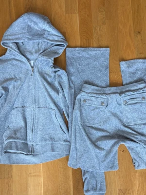 Grått loungewear-set Juicy Couture - Mysigt grått loungewear-set från Juicy Couture med zip-hoodie och matchande byxor. Hoodien har dragkedja och fickor, byxorna har bakfickor med knappar. Perfekt för chill dagar hemma. Mjuk och skön kvalitet.