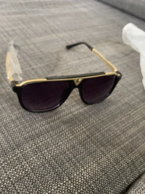Svarta solglasögon med gulddetaljer - Snygga solglasögon med svart båge och guldfärgade detaljer. Linsformen är klassisk aviator med mörka glas. Perfekt accessoar för dig som vill sticka ut med en trendig look.