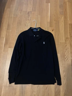 Svart långärmad polotopp från Polo Ralph Lauren - Klassisk svart långärmad polotopp från Polo Ralph Lauren med vit broderad logga på bröstet. Tidlös design med krage och knappar framtill. Perfekt för en clean och stilren look.