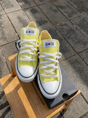 Gula Converse All Star sneakers  -  Snygga gula Converse All Star sneakers med klassisk låg modell. Skorna har vit tåhätta i gummi, vita snören och sula samt tydlig All Star logga på plösen. Ovandelen är i canvas och sulan är i gummi. Perfekta för en avslappnad streetstil. De är helt oanvända men ej nyköpta och säljs nu. Original kartongen finns kvar.