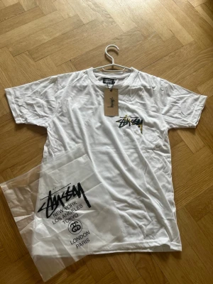 Vit Stüssy t-shirt med camo-logga - Säljer en fräsch vit t-shirt från Stüssy med deras ikoniska logga i camo-tryck på bröstet. Klassisk passform och rund hals. Perfekt till streetwear-stilen och kommer med originalpåse och tag kvar. Den passar folk med längden max 170cm ungefär men kontakt mig ifall ni har några funderingar runt detta!