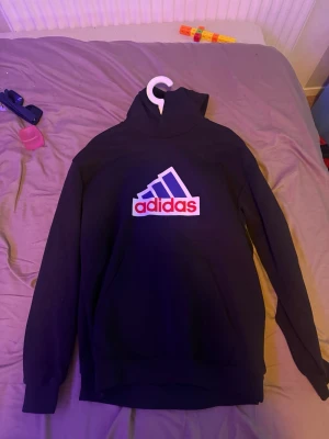 Svart hoodie från Adidas - Snygg svart hoodie från Adidas med stor logga i vitt, blått och rött på bröstet. Klassisk huva och ribbade muddar vid ärmslut och nederkant. Perfekt för en sportig och avslappnad stil.