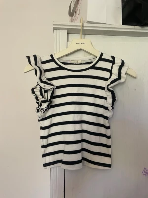 Randig volangtopp från Vero Moda - Säljer en vit och svart randig topp från Vero Moda med volangdetaljer vid ärmhålen. Toppen är ärmlös och har en rund halsringning. Materialet är ribbad bomull som ger en skön och stretchig känsla. Perfekt för dig som gillar en trendig och lekfull stil.