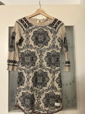 Mönstrad beige klänning Odd Molly - Unik beige klänning från Odd Molly med svart broderat mönster och dekorativa detaljer i rött och blått på axlar och ärmslut. Klänningen har trekvartsärm, rund halsringning med knyt och är tillverkad i mjuk bomull. Perfekt för dig som gillar bohemisk stil!