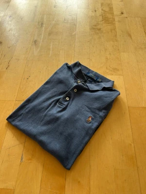Blå pikétröja från Polo Ralph Lauren - Klassisk blå pikétröja från Polo Ralph Lauren med vit knappslå och den ikoniska broderade loggan på bröstet. Tröjan har krage och är tillverkad i mjuk bomull som känns skön mot huden. Perfekt till jeans eller shorts för en avslappnad look. Äkta!