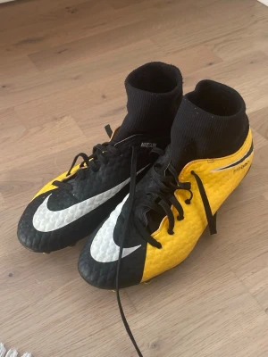 Nike Hypervenom svart/gul fotbollssko utomhus  - Säljer ett par Nike Hypervenom fotbollsskor i svart och gult med vita detaljer. Skorna har högt strumpskaft för extra stöd, snörning och en strukturerad ovandel i syntetmaterial för bättre bollkontroll. Perfekta för dig som vill sticka ut på planen!