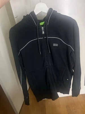 Svart hoodie från BOSS med dragkedja - Snygg svart hoodie från BOSS med vit piping längs bröstet och armarna. Hoodien har dragkedja framtill, två fickor och justerbar huva med snören. Liten BOSS-logga på bröstet. Perfekt för en avslappnad och stilren look.