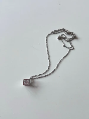 Silverhalsband med kubformad berlock - Ett silvrigt halsband med en liten ”kub” med en sten i. Sparsamt använt. Kuben är lite mindre än 1cm.  Kommer ej ihåg var det är köpt.