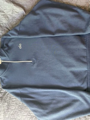 Blå half-zip hoodie från Alo - Mörkblå hoodie från Alo med half-zip dragkedja framtill och diskret logga på bröstet. Tillverkad i mjukt sweatshirtmaterial och har ribbade muddar vid ärmslut och nederkant. Perfekt för en chill och sportig look.