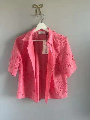 Rosa broderad blus Holly & Whyte - Söt rosa blus från Holly & Whyte med broderade blommor och hålmönster. Blusen har klassisk krage, korta ärmar med vågig kant och knäppning framtill. Tillverkad i bomull för en luftig och bekväm känsla. Perfekt för en trendig och somrig look. Min mamma köpte denna men den kom aldrig till användning. Världens finaste färg och tyg. 