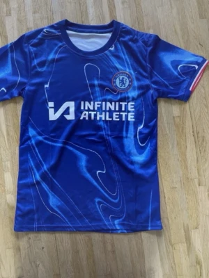 Chelsea FC blå matchtröja - Snygg blå Chelsea FC matchtröja med vita och ljusblå mönster, officiell logga på bröstet och sponsortryck 'INFINITE ATHLETE' framtill. Rund halsringning och korta ärmar med röd detalj på ärmslutet. Perfekt för fotbollsträning eller supporterliv.