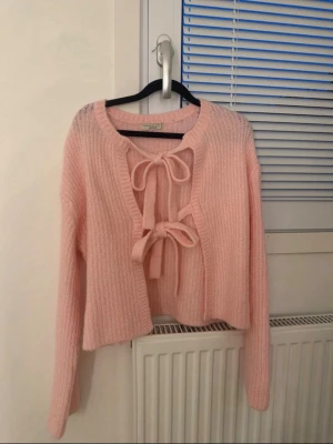 Ljusrosa stickad cardigan med rosetter - Supersöt ljusrosa stickad cardigan med två stora rosetter framtill. Tröjan har lång ärm, ribbad struktur och en croppad passform. Perfekt för att addera en gullig touch till din outfit.