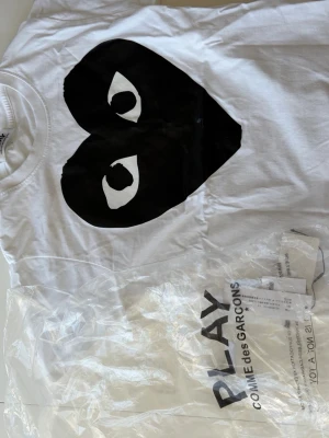 Vit Comme des Garçons Play t-shirt - Säljer en vit t-shirt från Comme des Garçons Play med det svarta hjärtat på bröstet. T-shirten har rund hals och korta ärmar. Perfekt för dig som gillar stilrena prints. Pris 350
