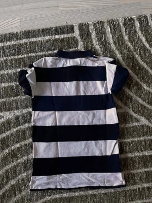 Randig pikétröja från Polo Ralph Lauren - Säljer en klassisk pikétröja från Polo Ralph Lauren med breda marinblå och vita ränder. Tröjan har korta ärmar, krage och knappar vid halsen. Perfekt för en avslappnad men ändå stilren look.