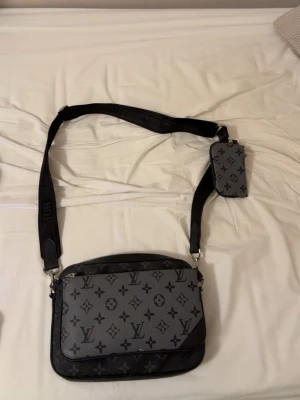 Louis Vuitton axelväska monogram grå - Säljer en snygg axelväska från Louis Vuitton i grått och svart med klassiskt monogrammönster. Väskan har justerbar axelrem och en liten avtagbar ficka. Tillverkad i slitstarkt läder och textil, perfekt för dig som vill ha en stilren och trendig accessoar. Priset kan diskuteras 
