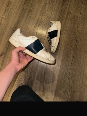 Vita sneakers med blå detalj - Snygga vita sneakers från Valentino med en bred mörkblå rem över mitten. Skorna har en klassisk låg profil och är tillverkade i läder med gummisula. Perfekta för dig som gillar stilrena och exklusiva sneakers.