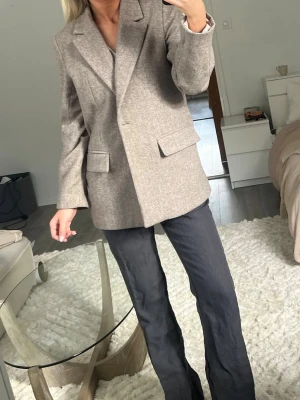 Beige kavaj från Zara - Stilren beige kavaj från Zara med klassisk krage och två fickor framtill. Kavajen har en rak passform och är tillverkad i ett mjukt, vävt material med diskret struktur. Perfekt för en trendig och avslappnad look. Ullkavaj