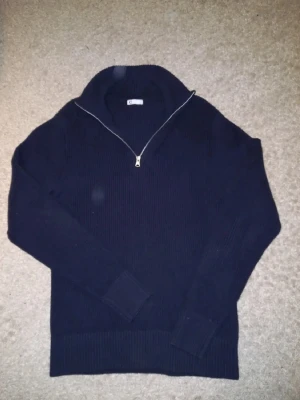 Mörkblå stickad tröja med dragkedja - Säljer en mörkblå stickad tröja med hög krage och dragkedja framtill. Tröjan har långa ärmar och ribbade muddar vid ärmslut och nederkant. Perfekt för lager på lager och en stilren look.