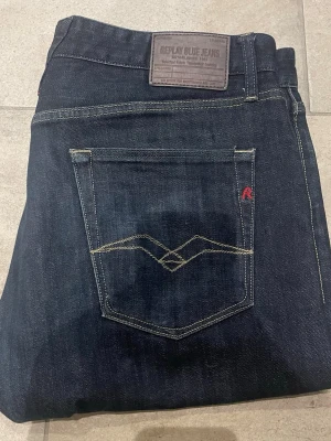 Mörkblå jeans från Replay - Snygga mörkblå jeans från Replay med klassisk femficksmodell och raka ben. Jeansen har kontrastsömmar, läderpatch bak i midjan och diskret röd detalj på bakfickan. Tillverkade i slitstark denim som ger en tidlös look. 34/36