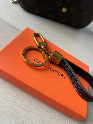 Vuitton nyckelring med läderdetalj - Snygg nyckelring från Vuitton med guldfärgad metall och brun flätad läderrem. Detaljer med LV-logga på både metall och läder. Perfekt accessoar för att hålla koll på nycklar eller hänga på väskan.