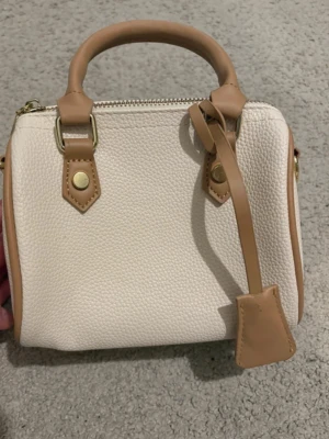 minihandväska - Snygg beige minihandväska med bruna detaljer och handtag. Väskan har guldfärgade detaljer och dragkedja upptill. Perfekt för det viktigaste och ger en clean look till din outfit.