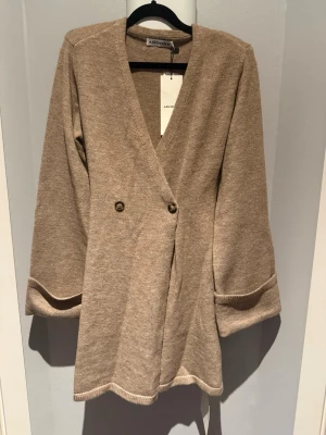 Beige omlottklänning från Adoore - Trendig beige omlottklänning från Adoore med v-ringning och dekorativa knappar framtill. Klänningen har långa ärmar med breda muddar och axelvaddar och är tillverkad i mjukt stickat material. Aldrig använd med prislappen kvar.