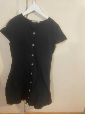 Svart kortklänning med knappar - Det är en 145-152, men jag tycker den är en 152-164. En svart kortklänning med korta ärmar och dekorativa knappar framtill. Klänningen har en enkel och stilren design som passar perfekt till sommaren. Tillverkad i ett mjukt tyg som känns bekvämt mot huden.