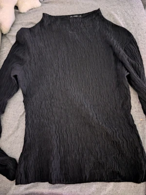 Svart långärmad topp från Gina Tricot stl xs - Säljer en svart långärmad topp från Gina Tricot i storlek XS. Toppen har ett vågigt, ribbat mönster i tyget som ger en cool struktur. Perfekt att styla med jeans eller kjol för en trendig look.