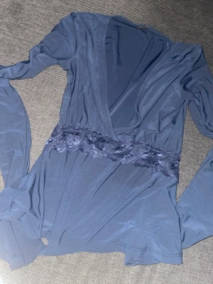 Marinblå tröja med lace runt midjan - Trendig blå topp med djup v-ringning och långa ärmar. Midjan har en bred spetsdetalj med blommönster som ger en extra snygg touch. Tillverkad i mjukt, stretchigt material som sitter skönt på kroppen. Har ett pyte litet hål vid bysten, men är inte synlig alls när man har på sig tröjan. Storlek är XXS, skulle passa XS. Säljer på grund av att den inte kommer till användning.