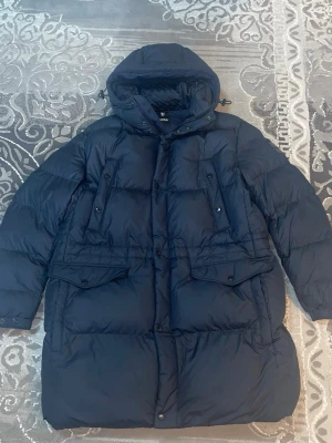 Mörkblå dunjacka från BOSS - Snygg mörkblå dunjacka från BOSS med stor huva och flera praktiska fickor framtill. Jackan har quiltad design, dragkedja och knappar samt justerbar huva. Perfekt för kalla vinterdagar och riktigt varm.