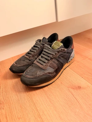 Valentino Garavani svarta sneakers - Snygga svarta sneakers från Valentino Garavani med detaljer i mocka och textil. Skorna har en klassisk låg silhuett, svarta skosnören och en beige sula med coola mönsterdetaljer. Perfekta för dig som vill ha en stilren men ändå unik look.