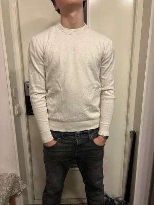 Gräddfärgad tjocktröja - Säljer en stilren gräddfärgad stickad tröja med rund halsringning och långa ärmar. Tröjan har en smal passform och ribbade muddar vid ärmslut och nederkant. Perfekt för en clean och enkel look.