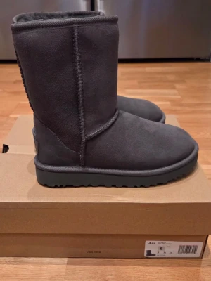 Mörkgrå UGG Classic Short boots - Mörkgrå UGG Classic Short boots i mocka med mjukt foder på insidan. Klassisk rund tå och grov sula för extra grepp. Perfekta för kalla dagar och riktigt sköna att ha på sig. Tidlös design med UGG-logga bak på hälen.
