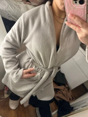 Ljusgrå fluffig morgonrock - Mysig ljusgrå morgonrock i supermjukt fleece-material med knytskärp i midjan och långa ärmar. Perfekt för att chilla hemma och hålla sig varm. Enkel och stilren design utan synligt märke.