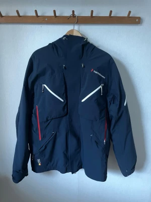 Mörkblå vindjacka från Peak Performance - Säljer en klassisk skaljacka från Peak Performance i toppskick! Modellen tillhör den tekniska Heli-serien (Heli Gravity) och är känd för sin grymma kvalitet och hållbarhet.  • Storlek: Medium (Herr) • Skick: Mycket bra, inga hål eller fläckar. • Färg: Marinblå med vita och röda detaljer. • Funktioner: Recco-reflektor, snölås, hjälmanpassad huva, ventilationsdragkedjor och putsduk för goggles. Perfekt för både offpist och i backen. Vind- och vattentät med grym passform.