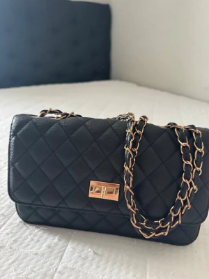 Svart quiltad handväska med kedja - Snygg svart handväska i quiltad design med guldfärgad kedjerem och vridlås. Klassisk rektangulär form och elegant look som passar till många outfits. Väskan har en lyxig känsla tack vare materialet och detaljerna.