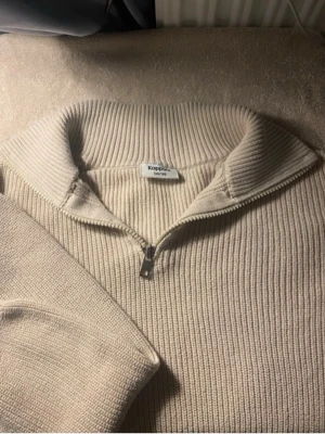 Beige stickad halvzip från Kappahl - Snygg beige stickad tröja från Kappahl med halvzip och hög krage. Tröjan har ribbstickad struktur och breda muddar vid ärmslut och nederkant. Perfekt för lager på lager och enkel att matcha med jeans eller byxor.