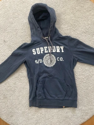 Mörkblå hoodie från Superdry - Mörkblå hoodie från Superdry med stor vit logga och broderad detalj på bröstet. Klassisk känguruficka framtill och justerbar huva med snören. Tillverkad i mjukt bomullsmaterial, perfekt för en avslappnad och sportig stil. Passar S bra.