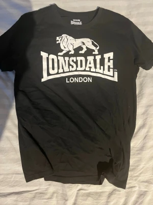 Svart Lonsdale t-shirt  - Säljer en svart t-shirt från Lonsdale med stort vitt tryck av loggan och ett lejon på bröstet. Klassisk rund hals och korta ärmar. Perfekt för dig som gillar streetwear och vill ha en enkel men snygg look.