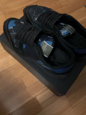 Valentino blå camo sneakers - Snygga sneakers från Valentino med blått och svart camo-mönster, svarta skosnören och detaljer i mocka och textil. Perfekt inför våren