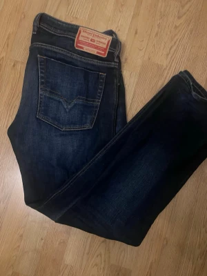 Mörkblå jeans från Diesel Industry - Snygga mörkblå jeans från Diesel Industry med klassisk femficksdesign och raka ben. Jeansen har tydliga kontrastsömmar och en läderpatch med logga bak i midjan. Perfekta för dig som gillar en tidlös och cool look. Storleken är 36/32 men de sitter mer som 36/34 är 190 och det sitter helt perfekt i längden